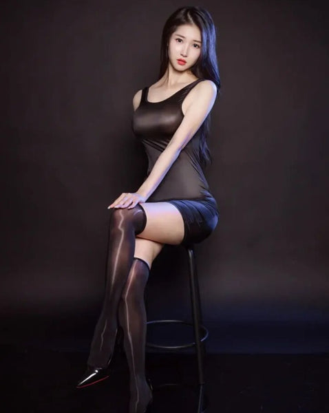 A woman posing in a black glossy mini dress with thick shoulder straps, a scoop neckline, and a low cut back, with black thigh high stockings and black high heels.